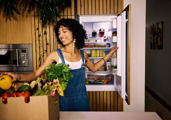 Vrouw staat in de keuken en doet de koelkast open met een lach