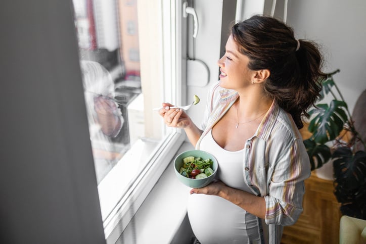 Een zwangere vrouw eet een gezonde salade terwijl ze uit het raam kijkt en glimlacht.