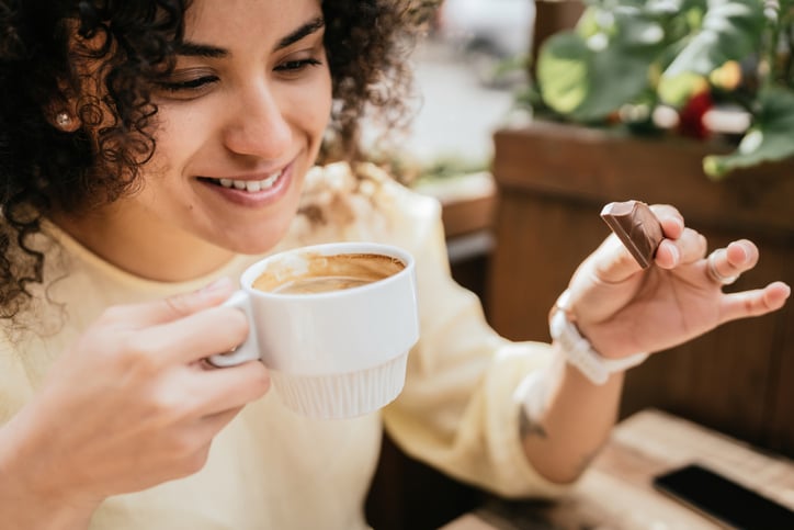 Vrouw kijkt lachend naar kopje koffie met een stukje chocola in haar hand.